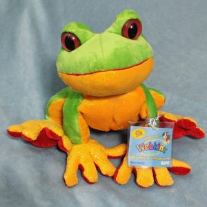 May include: A plush frog toy with a green head and back, orange belly and legs, and red accents. The frog has large red eyes and a tag that reads "Webkinz". The toy is made of soft, fuzzy material.