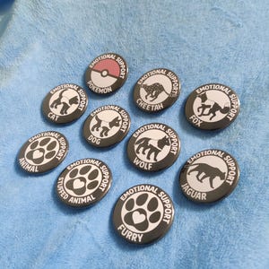 May include: A selection of black and white enamel badges, each bearing the words "Emotional Support" and an animal illustration, such as a cat, dog, or wolf. Other designs include a Pokemon ball, paw prints, and the word "Furry".