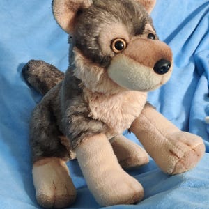 May include: A plush wolf toy with a realistic design. The toy features a mix of gray, brown, and beige fur, with a detailed face and expressive eyes. The wolf is posed in a playful stance, with its paws extended. The background is a solid blue.