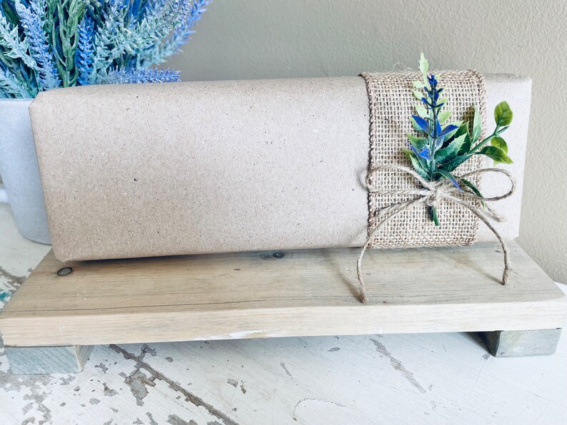 May include: A brown paper-wrapped gift box with a burlap bow and blue and green floral accents. The box is sitting on a wooden stand.