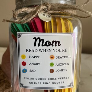 May include: A glass jar filled with colourful paper slips, tied with twine and a small metal tag that says "Love". The jar has a label that reads "Mom Read When You're" with a colour-coded key for different emotions, and "Color Coded Bible Verses 60 Inspiring Quotes".