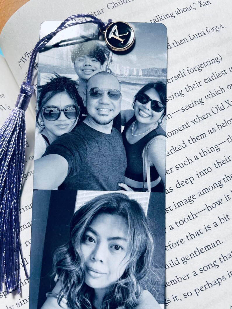 May include: A purple tassel bookmark with a metal charm featuring the letter K. The bookmark has two photos of a family. The top photo shows a family of four, and the bottom photo shows a woman with long dark hair.