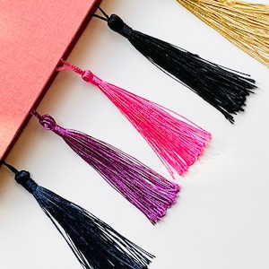 May include: Five colorful tassel bookmarks attached to a red book. The tassels are gold, black, pink, purple, and dark blue.
