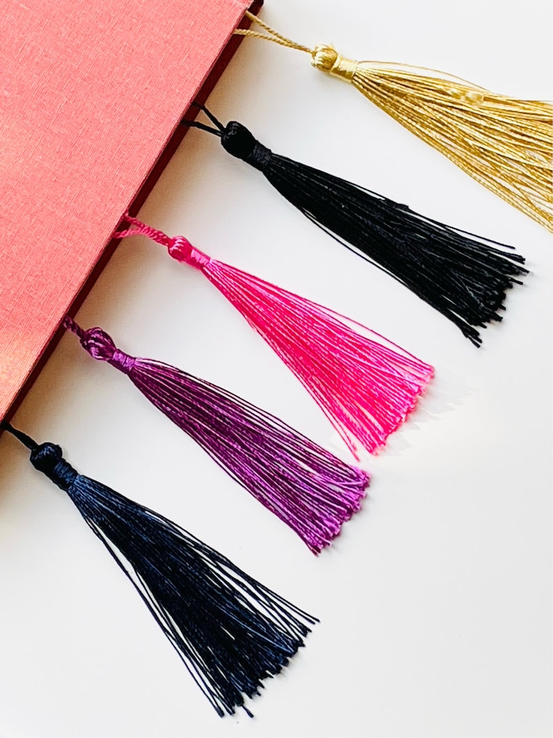 May include: Five colorful tassel bookmarks attached to a red book. The tassels are gold, black, pink, purple, and dark blue.