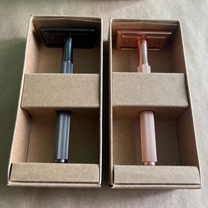 May include: Two safety razors in cardboard boxes. One razor is black with a silver handle, and the other is rose gold with a rose gold handle. The razors are in a brown cardboard box with the words "RECYCLE ME" printed on the lid.