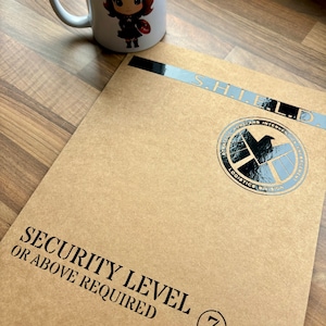 May include: A brown cardboard folder with the text "S.H.I.E.L.D." and "SECURITY LEVEL 7 OR ABOVE REQUIRED" printed on it. The folder has a black and silver S.H.I.E.L.D. logo sticker on the front.