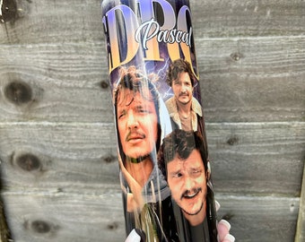 May include: A white tumbler with a black and white image of Pedro Pascal. The image is a collage of three different photos of Pascal. The tumbler has a black background with a purple and yellow gradient at the top. The text "Pedro Pascal" is written in white cursive on the tumbler.