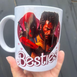 May include: White ceramic mug with a red heart design featuring Deadpool and Wolverine. The heart is surrounded by pink hearts. The text "Bestie" is printed in red letters on the mug.