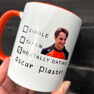 May include: White ceramic mug with an orange rim and handle. The mug has a checklist with the options "Single", "Taken", and "Mentally Dating Oscar Piastri" checked. A photo of Oscar Piastri is in the background.