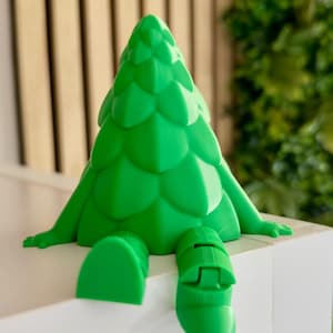 May include: A vibrant green decorative tree figurine with a whimsical design. The tree has arms and legs, appearing to sit on a white surface. The background includes a wooden slatted wall and some greenery. The figurine is a fun, modern design.