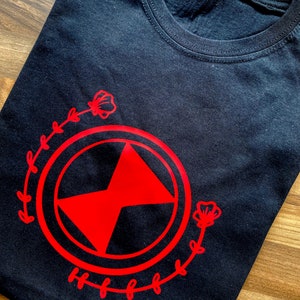 May include: Black t-shirt featuring a red graphic design of a stylized symbol encircled by a wreath of leaves and flowers.