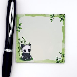 May include: A black pen rests next to a square notepad with a panda illustration. The notepad features a green border with bamboo and leaf accents. The panda is black and white, with large eyes. The notepad has the text "Kyashi".