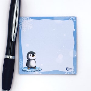 Könnte beinhalten: Ein blauer Notizblock mit einer Pinguin-Illustration und einem weißen Schneeflocken-Design. Der Notizblock trägt den Text "Kyashi" in einem Kreis unten.