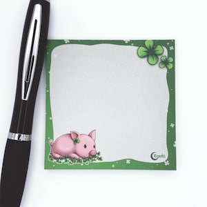 Könnte beinhalten: Grüner Notizblock mit einem rosa Schweinchen und Kleeblatt-Designs. Der Notizblock hat einen weißen Schreibbereich und den Text "Kyashi" unten.