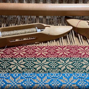 Pode incluir: Uma aproximação de um tear com duas lançadeiras de madeira. Uma lançadeira contém fio dourado, a outra fio vermelho. O tecido tecido exibe um padrão geométrico em vermelho, verde e azul.