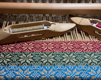 May include: Two light-coloured wooden weaving shuttles resting on a loom. One shuttle holds a metallic gold thread bobbin, while the other holds a red thread bobbin. The loom features a woven fabric with geometric patterns in red, green, and blue.