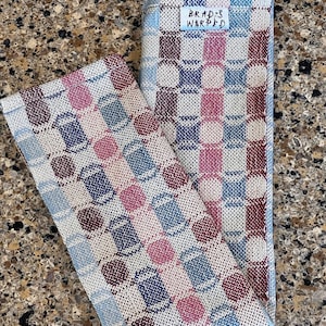 Op de afbeelding: Twee geweven theedoeken met een geometrisch patroon in tinten blauw, roze en bordeauxrood. De theedoeken zijn gevouwen en hebben een label met de tekst "Brad's Warped".
