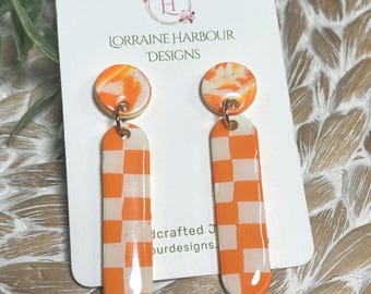 May include: Dangle earrings featuring a circular top piece with an orange and white marbled pattern and a long, rectangular bottom piece with an orange and white checkerboard pattern. The earrings are displayed on a white card labeled Lorraine Harbour Designs.