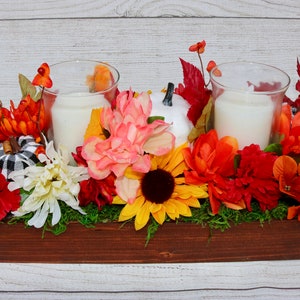 Puede incluir: Un centro de mesa de madera con temática otoñal. Presenta calabazas blancas, flores rojas, naranjas y amarillas, y dos candelabros de vidrio con velas blancas. El centro de mesa está rodeado de musgo verde.
