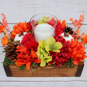Könnte beinhalten: Ein Holzkasten-Mittelstück mit einer weißen Kerze in einem Glashalter, umgeben von roten, orangefarbenen und grünen Kunstblumen, weißen Kürbissen und Tannenzapfen.