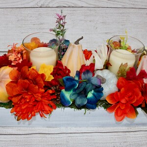 Könnte beinhalten: Ein weißer Holzkasten mit Herbstdekoration. Er enthält orange, rote und blaue Blumen, Kürbisse und zwei Glaskerzenhalter mit weißen Kerzen.