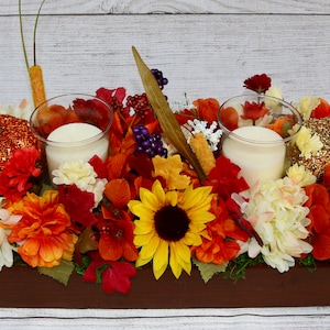 Könnte beinhalten: Ein Holzbox-Mittelstück mit Herbstdekoration. Es verfügt über zwei weiße Kerzen, künstliche Kürbisse, Sonnenblumen und andere Herbstlaub in Orange-, Rot- und Gelbtönen.