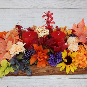 Könnte beinhalten: Ein Holz-Mittelstück mit Herbstdekorationen, darunter rote Kerzen, orange und gelbe Blumen und braune Blätter.
