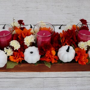 Könnte beinhalten: Ein Holz-Mittelstück mit drei roten Kerzen, weißen Kürbissen und orangefarbenen und roten Blumen.