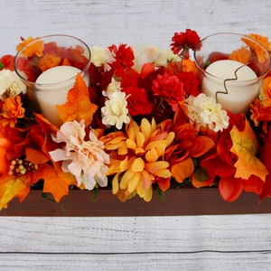 Könnte beinhalten: Ein Holz-Mittelstück mit Herbstdekoration, darunter orangefarbene Kürbisse, rote und orangefarbene Blumen und zwei Glaskerzenhalter mit weißen Kerzen.