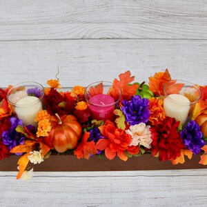 Könnte beinhalten: Ein Holzbox-Mittelstück, dekoriert mit Herbstlaub, Blumen und Kürbissen. Das Mittelstück enthält drei gläserne Kerzenhalter mit weißen und rosa Kerzen.
