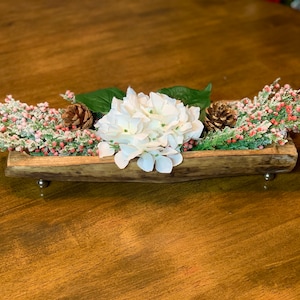 May include: A wooden trough with metal feet, filled with white hydrangeas, green sprigs, and red berries. The trough is on a wooden table.