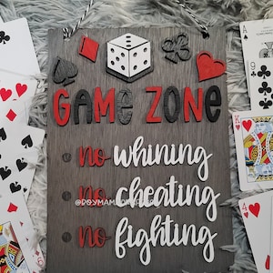 May include: A wooden sign with the words "GAME ZONE" in red and black letters. Below the words are three rules: "no whining", "no cheating", and "no fighting". The sign is decorated with playing card symbols, a dice, and a heart.