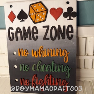 May include: A wooden sign with the words "Game Zone" in black letters on a white background. Below the words are three rules: "no whining", "no cheating", and "no fighting". The sign is decorated with a red diamond, a black spade, a yellow die, a red heart, and a black club.