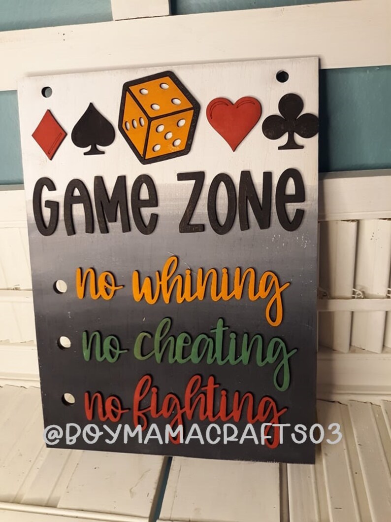 May include: A wooden sign with the words "Game Zone" in black letters on a white background. Below the words are three rules: "no whining", "no cheating", and "no fighting". The sign is decorated with a red diamond, a black spade, a yellow die, a red heart, and a black club.