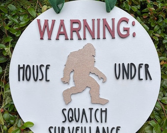 May include: White round wooden sign with the text "WARNING: HOUSE UNDER SQUATCH SURVEILLANCE" in black lettering. A brown silhouette of a Bigfoot is in the center of the sign. The sign is hanging from a green ribbon.