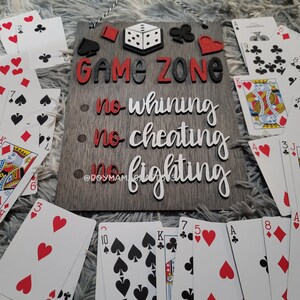 May include: A wooden sign with the words "GAME ZONE" in red and black letters. The sign also has the words "no whining", "no cheating", and "no fighting" written in white letters on a gray background. The sign is surrounded by playing cards.