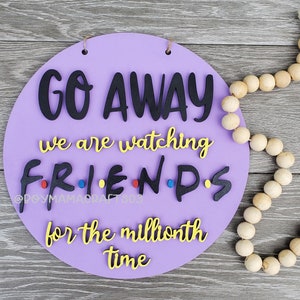 May include: A round wooden sign in purple with the text "Go Away we are watching FRIENDS for the millionth time" in black and yellow lettering. The word FRIENDS is spelled out with individual letters and has red, blue, and yellow dots between the letters.