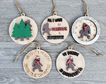 May include: Five wooden Christmas ornaments featuring a Bigfoot character in various poses. The ornaments are decorated with red, green, and black paint. The text on the ornaments reads: "All I want for Christmas is...