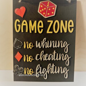 May include: A black wooden sign with the words "Game Zone" in yellow letters. Below the words are three rules: "no whining", "no cheating", and "no fighting". Each rule is accompanied by a playing card symbol: a spade, a diamond, and a club.