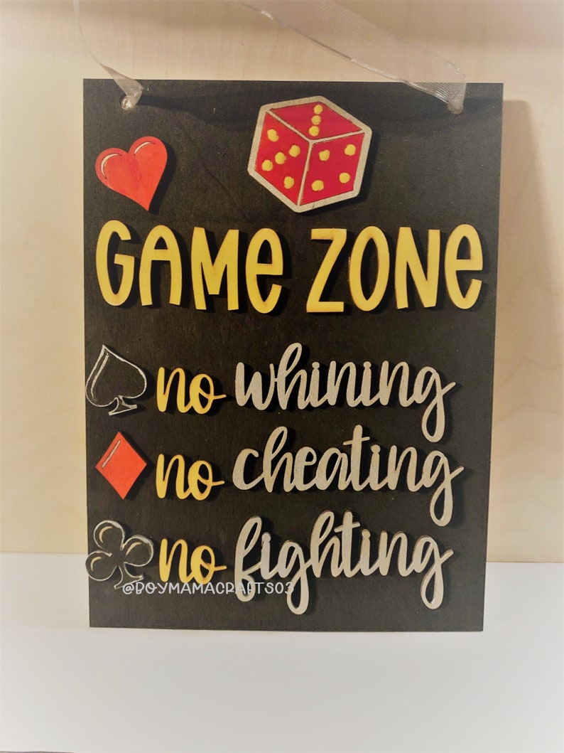 May include: A black wooden sign with the words "Game Zone" in yellow letters. Below the words are three rules: "no whining", "no cheating", and "no fighting". Each rule is accompanied by a playing card symbol: a spade, a diamond, and a club.