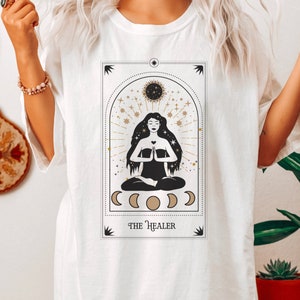 May include: White t-shirt with a graphic of a tarot card featuring a woman meditating with the text "THE HEALER" at the bottom.