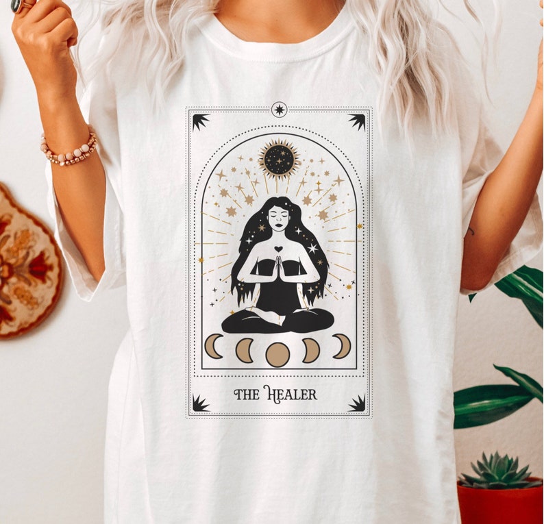 May include: White t-shirt with a graphic of a tarot card featuring a woman meditating with the text "THE HEALER" at the bottom.