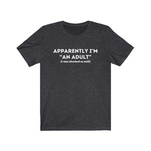 May include: A dark heather grey t-shirt with the text "Apparently I'm "An Adult" (I was shocked as well)" printed in white.