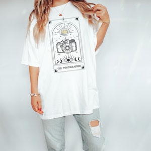 May include: White oversized t-shirt with a graphic of a vintage camera in a tarot card style frame. The text "THE PHOTOGRAPHER" is below the image.