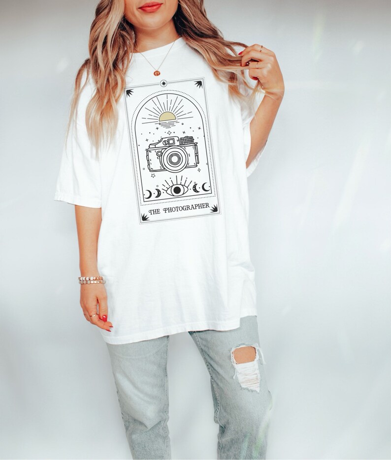 May include: White oversized t-shirt with a graphic of a vintage camera in a tarot card style frame. The text "THE PHOTOGRAPHER" is below the image.