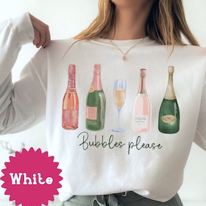 May include: White sweatshirt with a graphic of five champagne bottles and a champagne flute. The text "Bubbles please" is printed below the bottles.