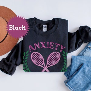 May include: Black sweatshirt with the text "ANXIETY" in pink lettering and two pink tennis rackets with green leaves on either side.