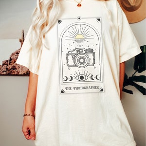 May include: A white oversized t-shirt with a black and yellow graphic of a camera, sun, moon, and eye. The text "THE PHOTOGRAPHER" is printed below the graphic.