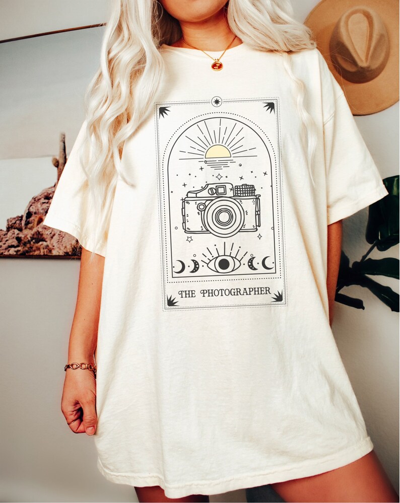 May include: A white oversized t-shirt with a black and yellow graphic of a camera, sun, moon, and eye. The text "THE PHOTOGRAPHER" is printed below the graphic.
