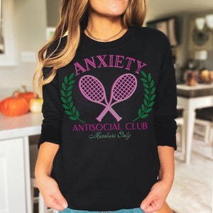May include: Black sweatshirt with a pink graphic of two crossed tennis rackets with green laurel wreaths and the text "Anxiety Antisocial Club Members Only".